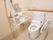 白い空間に、木目調の棚を合わせたトイレである。壁付の手すりと可動式の手すりと、手洗いボウルが設置されている。