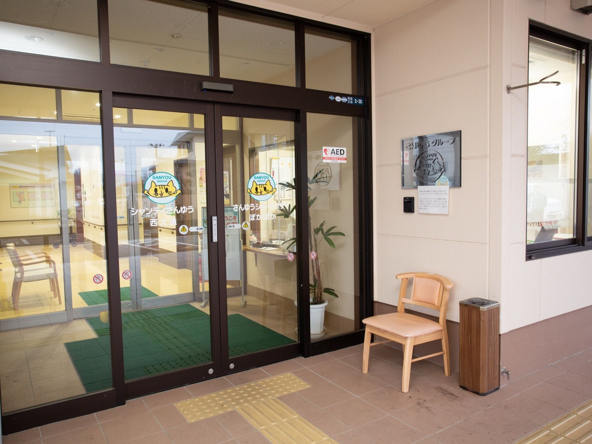 灰皿スタンドの隣に椅子が１脚置かれている。点字ブロックが設置されている。屋外用ドアマットが用意されている。