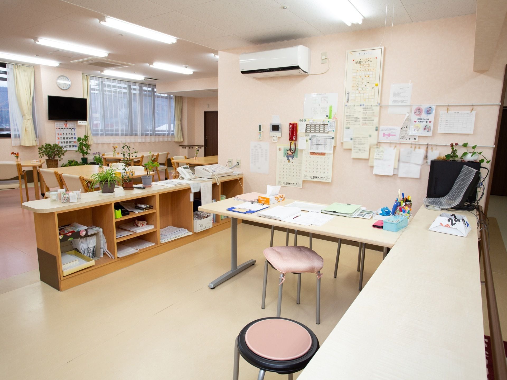 観葉植物がたくさん置かれている。エアコンとテレビが設置されている。壁にカレンダーや書類などが貼られている。