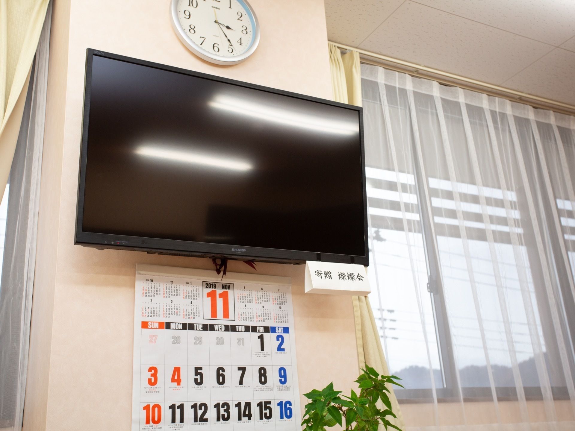 窓にはレースのカーテンが掛けられている。テレビが設置されており、壁にはカレンダー、時計が飾られている。