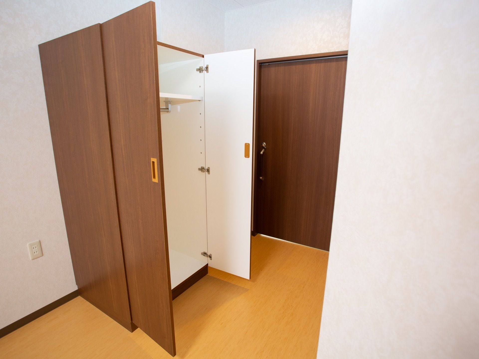 フローリングの居室で、白い壁になっている。床の近くにコンセントがある。壁際に観音開きのタンスが用意されている。