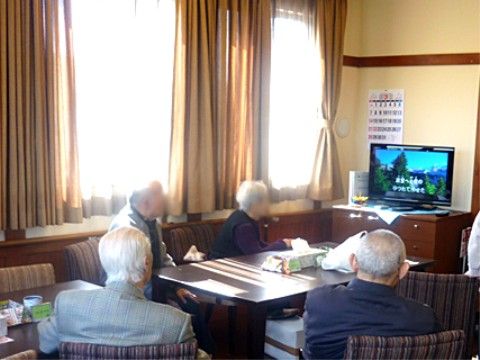 大きな窓から日差しが差し込んでいる。窓際に長テーブルやソファが置かれている。チェストの上にあるテレビにはカラオケの画面が映っている。