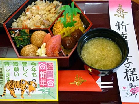 お盆に並べられたお正月の食事には新年の挨拶が添えられている。赤い割り箸袋には「壽」と印刷されている。