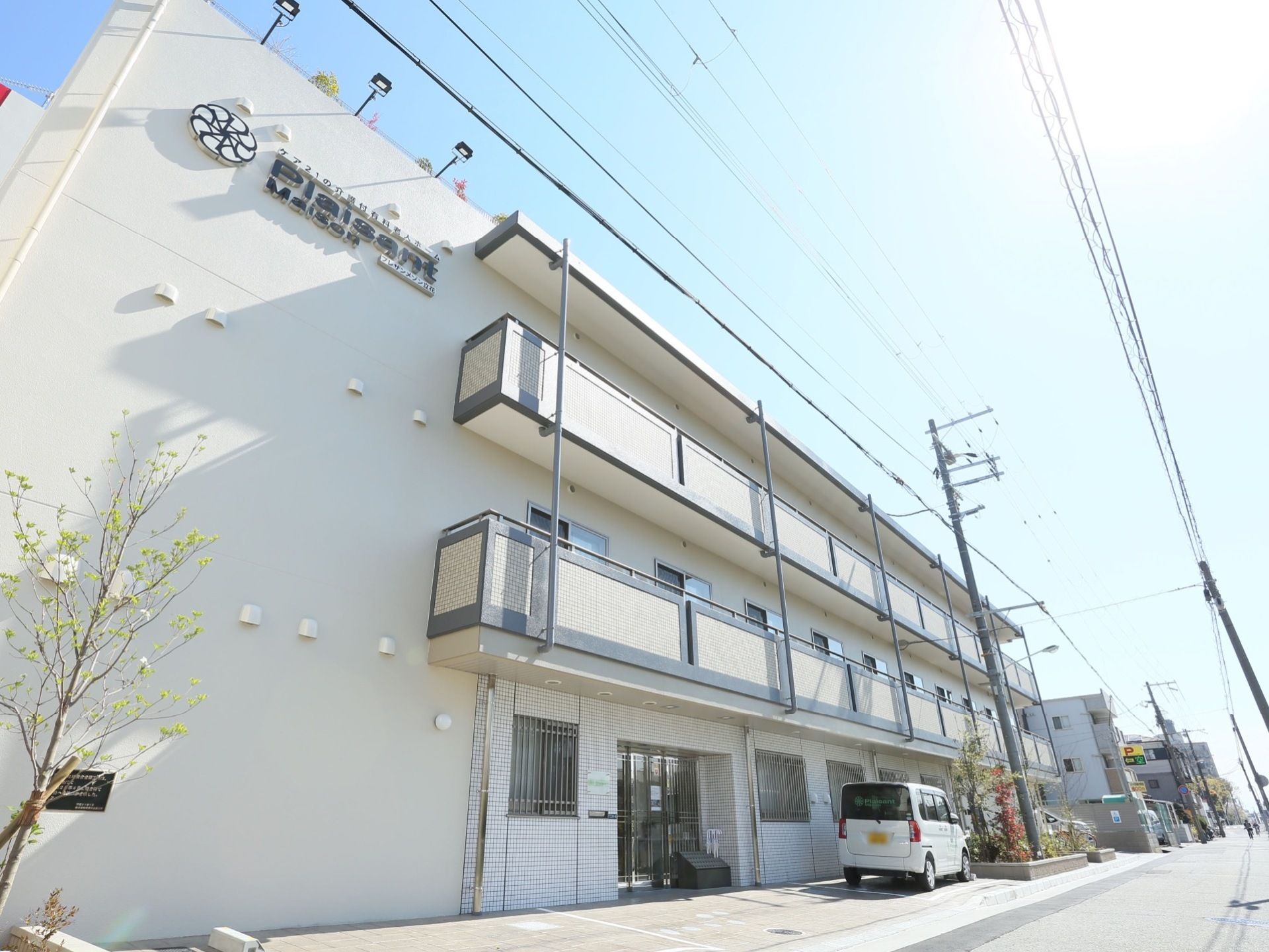 地上４階建ての施設は、マンションのような都会的な外観デザインである。左端には、枝の細い樹木が植わっている。
