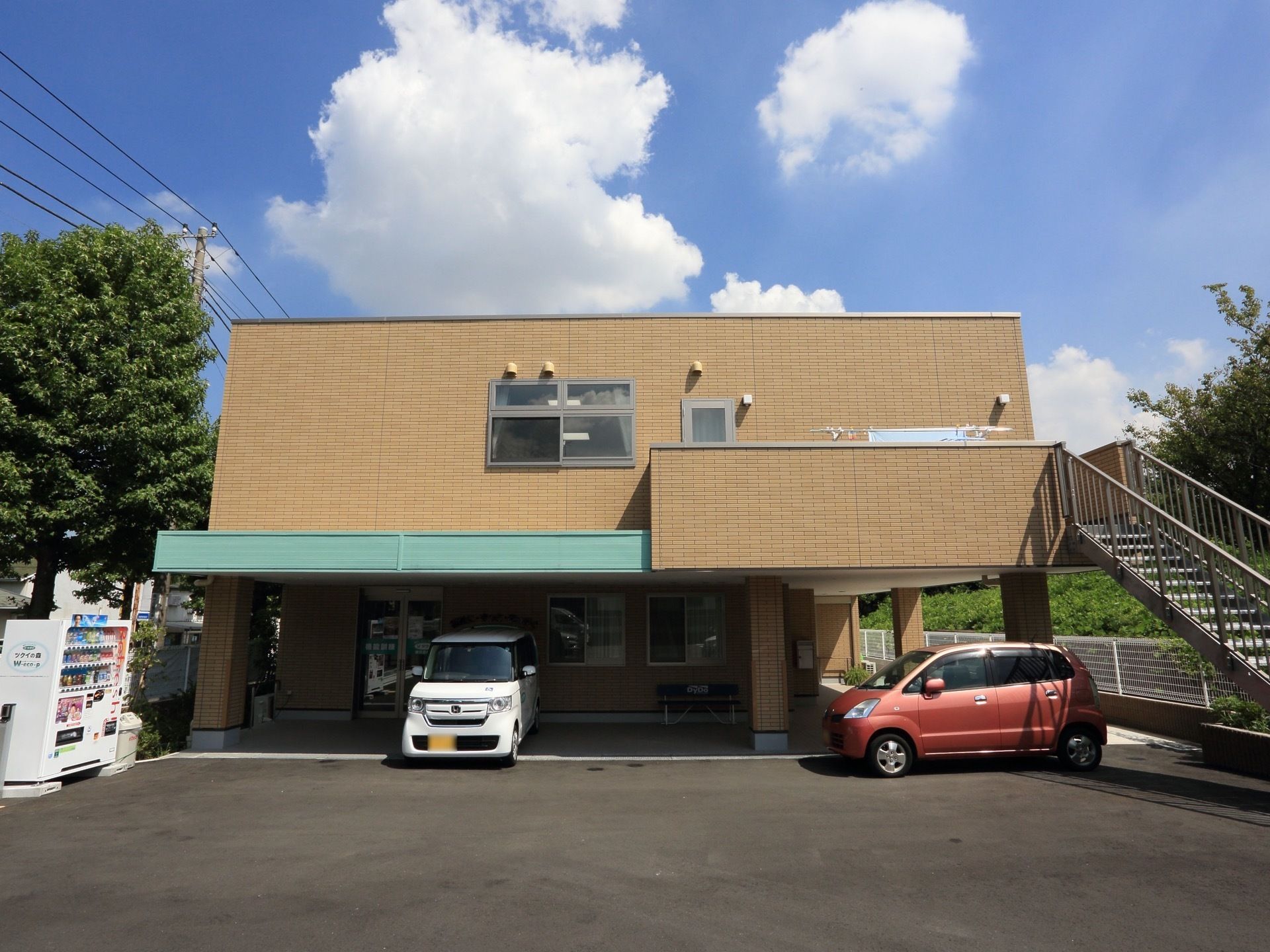 周辺環境に馴染む、落ち着いた色合いの建物である。敷地はアスファルトで広々としており、複数の車が駐車できる。