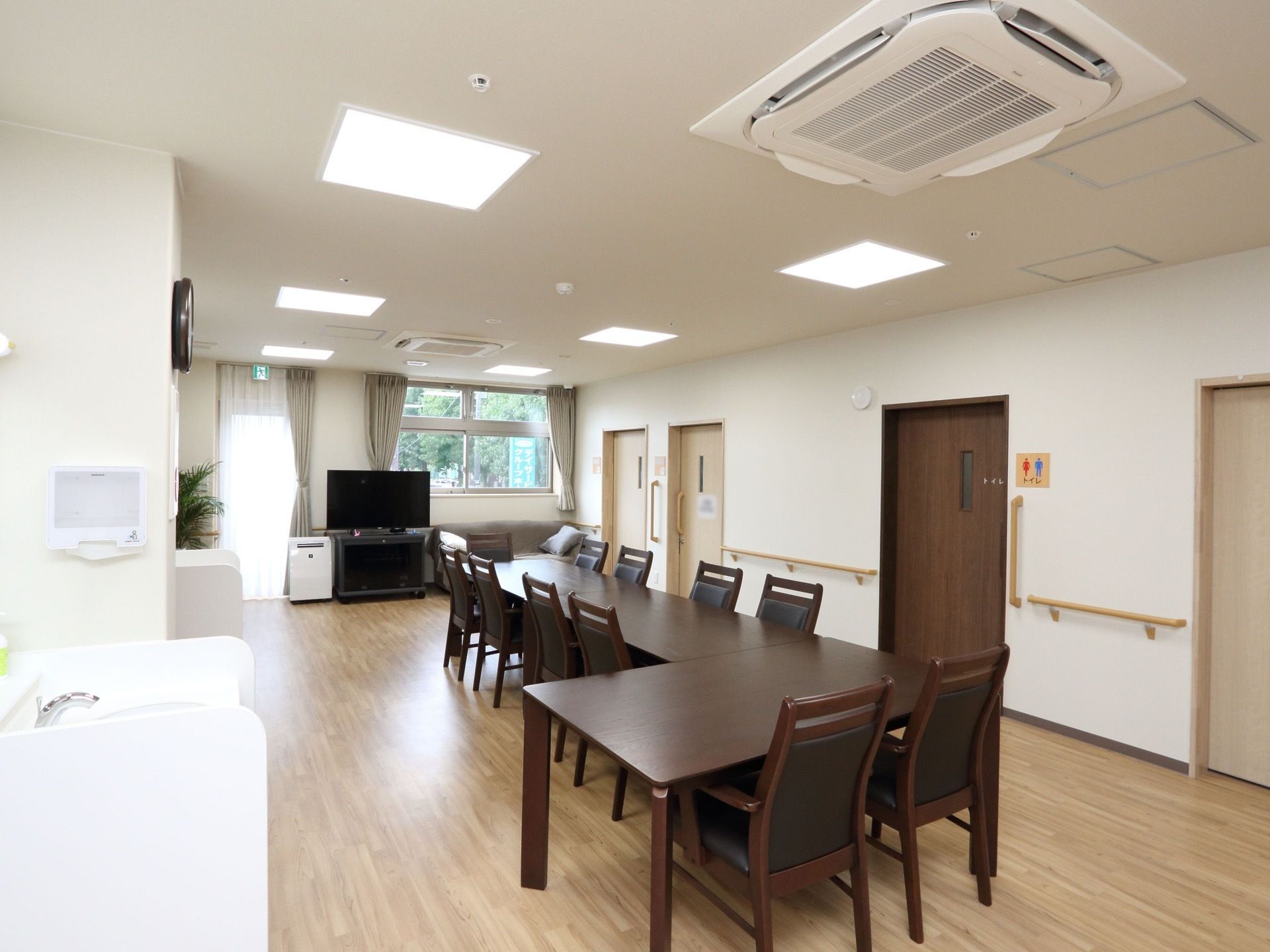 L字型にテーブルが配置され、椅子が並べられている。そばにはトイレや洗面台があり、窓の側にはテレビが置いてある。