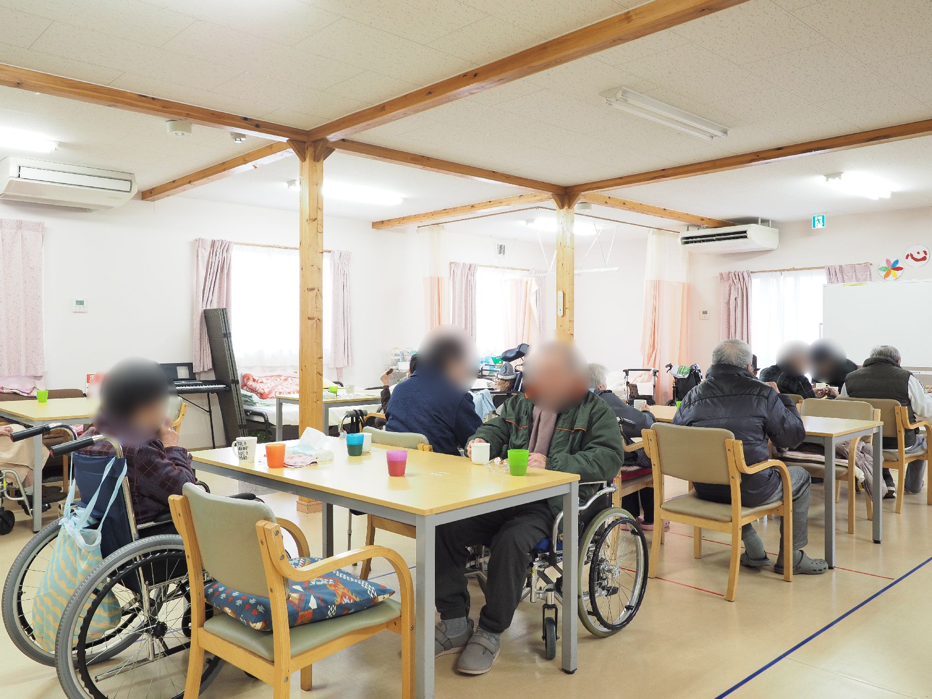 共有スペースに入居者様が集い、ダイニングテーブルを囲んでお茶を飲んだり、談話をしたりしている。窓にはピンク系のカーテンが吊り下がっている。