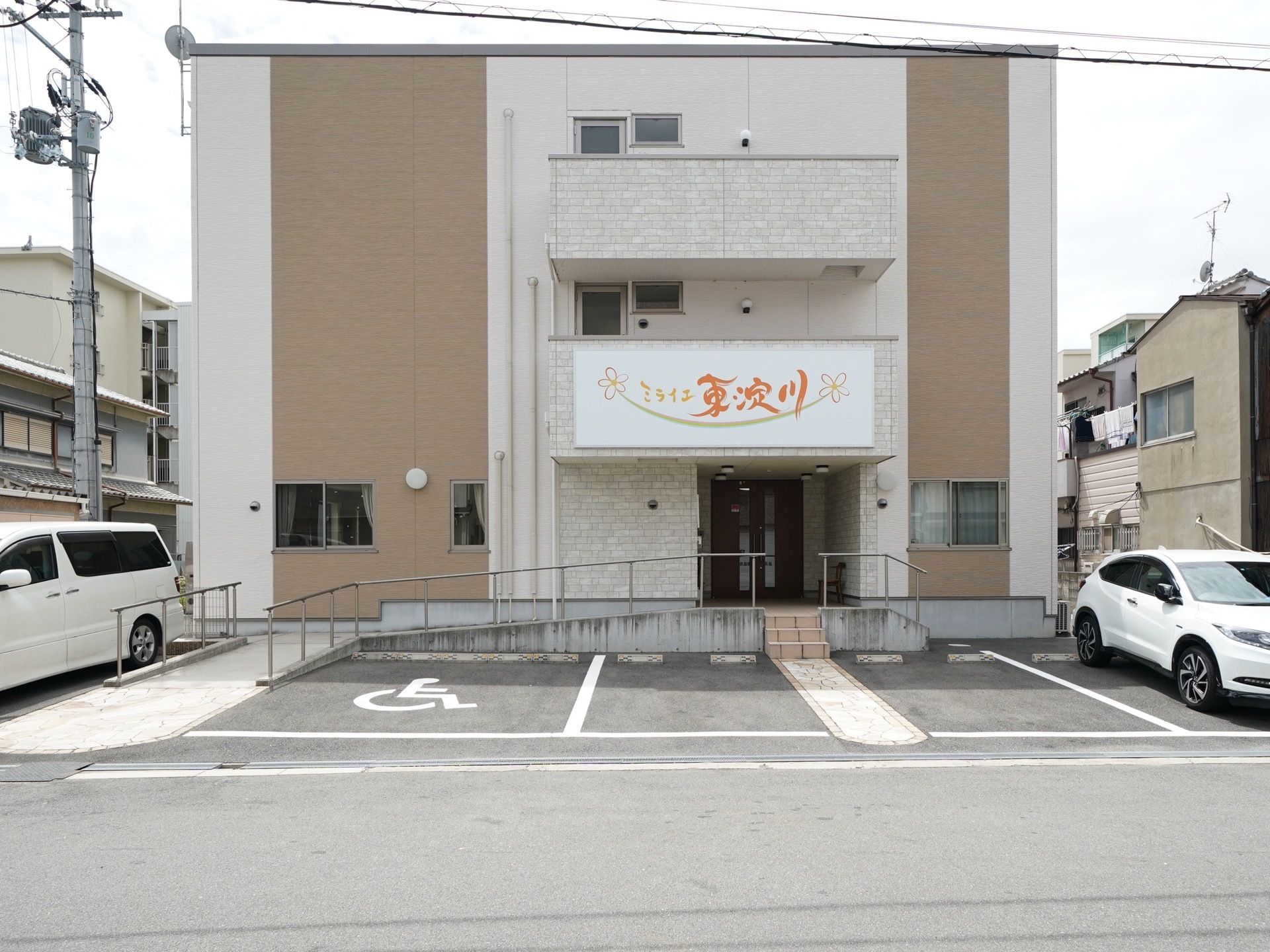 白色をベースとした建物に、オレンジ色の文字で施設名が書いてある看板が設置されている。アスファルトで舗装された駐車場がある。