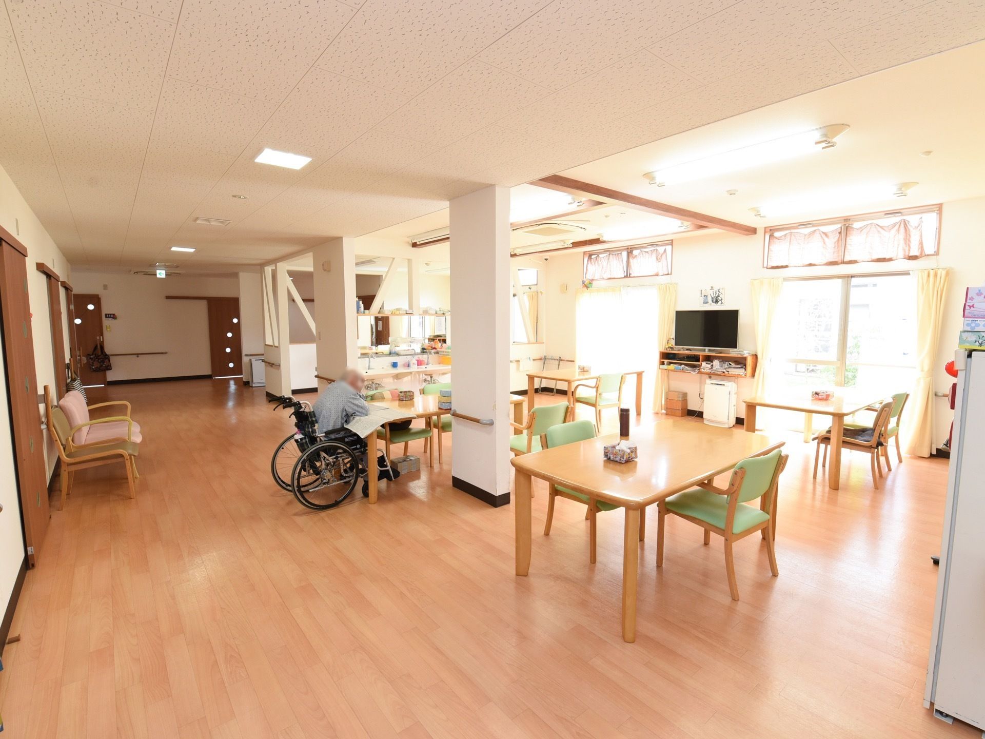 天井の高い、開放感あふれる食堂。居室の通路に併設している。大きな窓が複数あって明るい。テーブルと椅子がたくさん並んでいる。