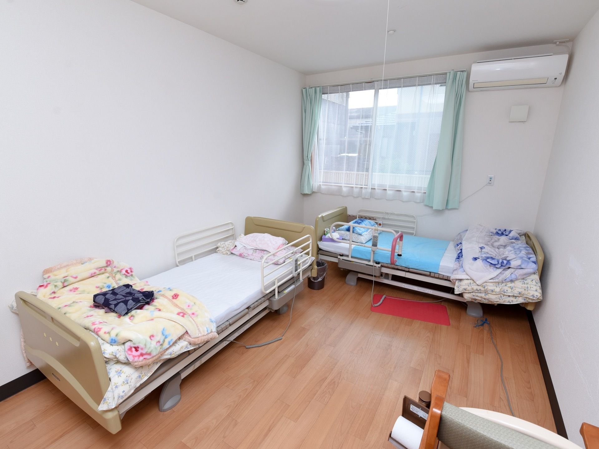 入居者様の居室に介護用ベッドが２つ置かれている。窓にはカーテンが掛けられ、壁にはエアコンが設置されている。