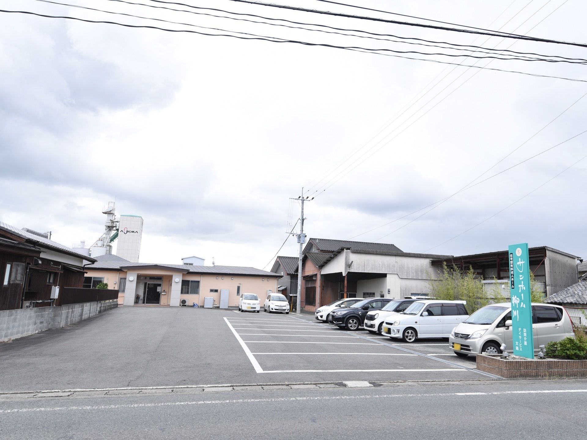 施設の手前が広い駐車場になっており、車が数台停まっている。入り口にはライトグリーンの大きな看板があり、施設名が書かれている。
