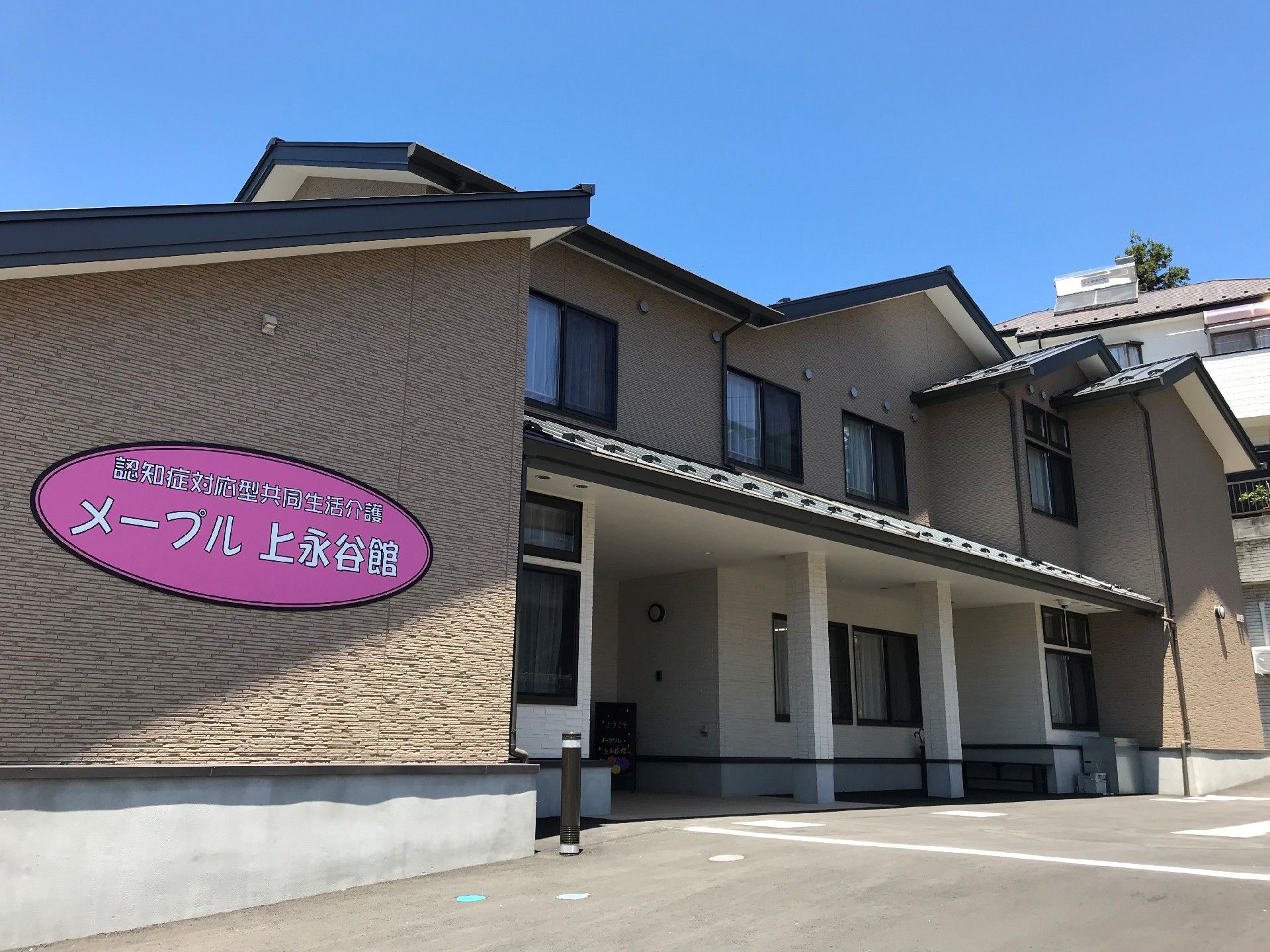 大きな一軒家のような建物を左手前から見た様子。2階建ての茶色を基調とした住まいで、壁にはピンク色の楕円形の中にホームの名前が書かれている。