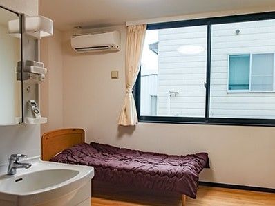 居室にはエアコンと洗面所があり、窓が大きく明るい雰囲気。ベッドには茶色の布団がセットされている。