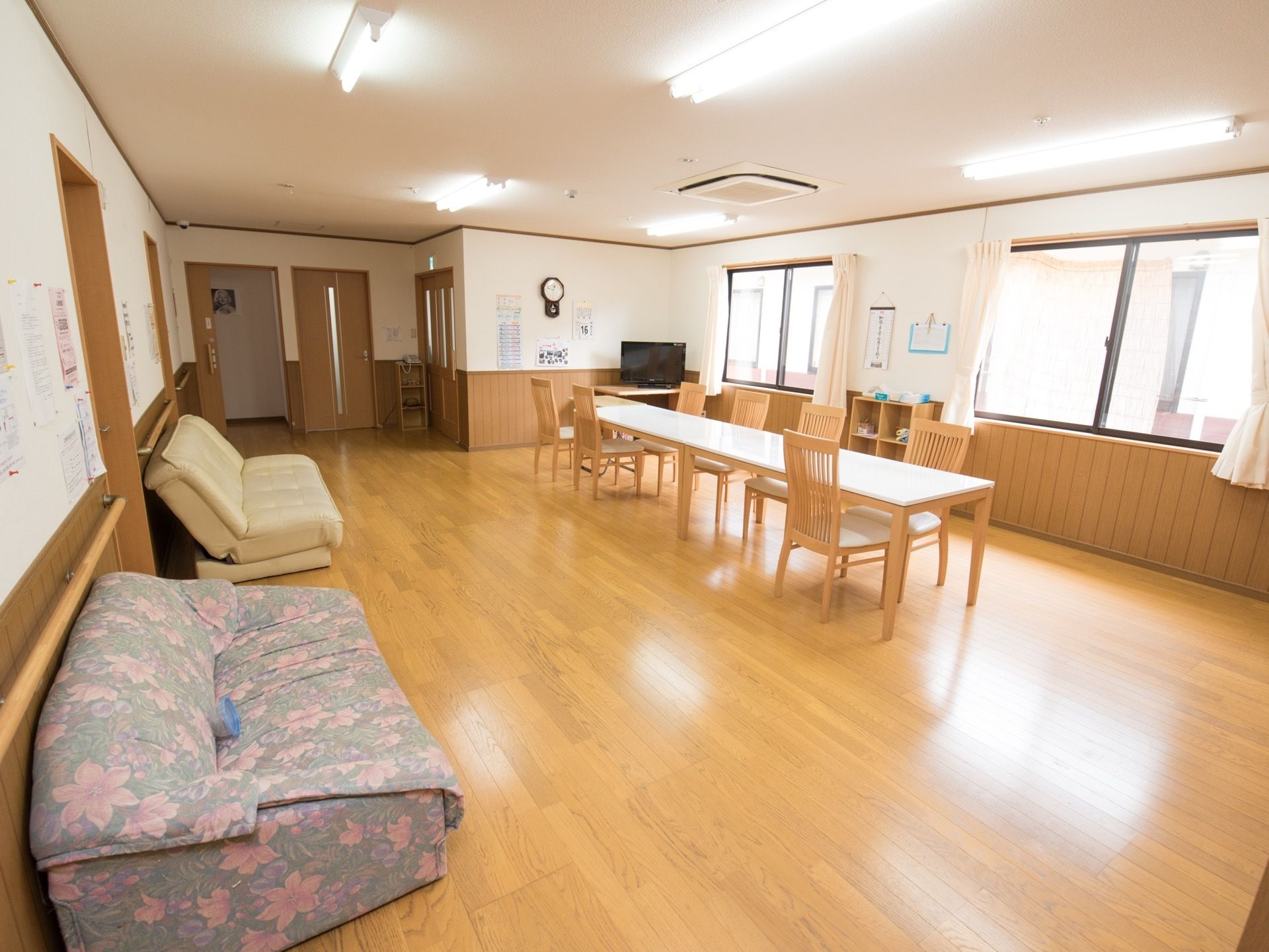 カーテンの掛かった窓が複数設置されており、天井のライトと合わせて明るい空間になっている。壁側にソファーが置かれている。