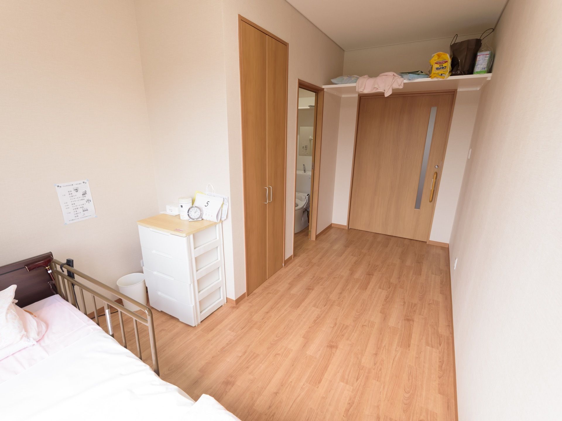 床や扉が淡い木目で統一されている明るい雰囲気の居室である。部屋の奥にベッドが置かれており、出入口近くにトイレが見える。