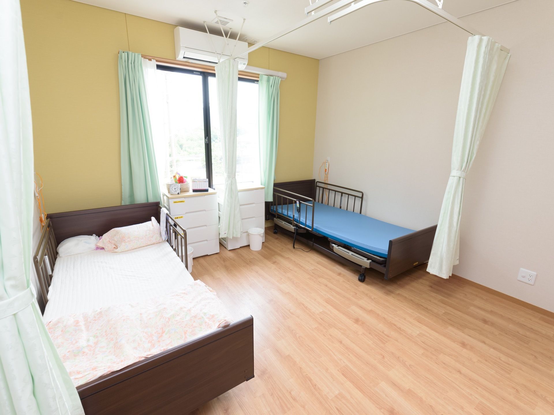 黄色い壁に窓が設けられている部屋にベッドが２台並んでいる。１台のベッドの周りにパーテーションカーテンが取り付けられている。