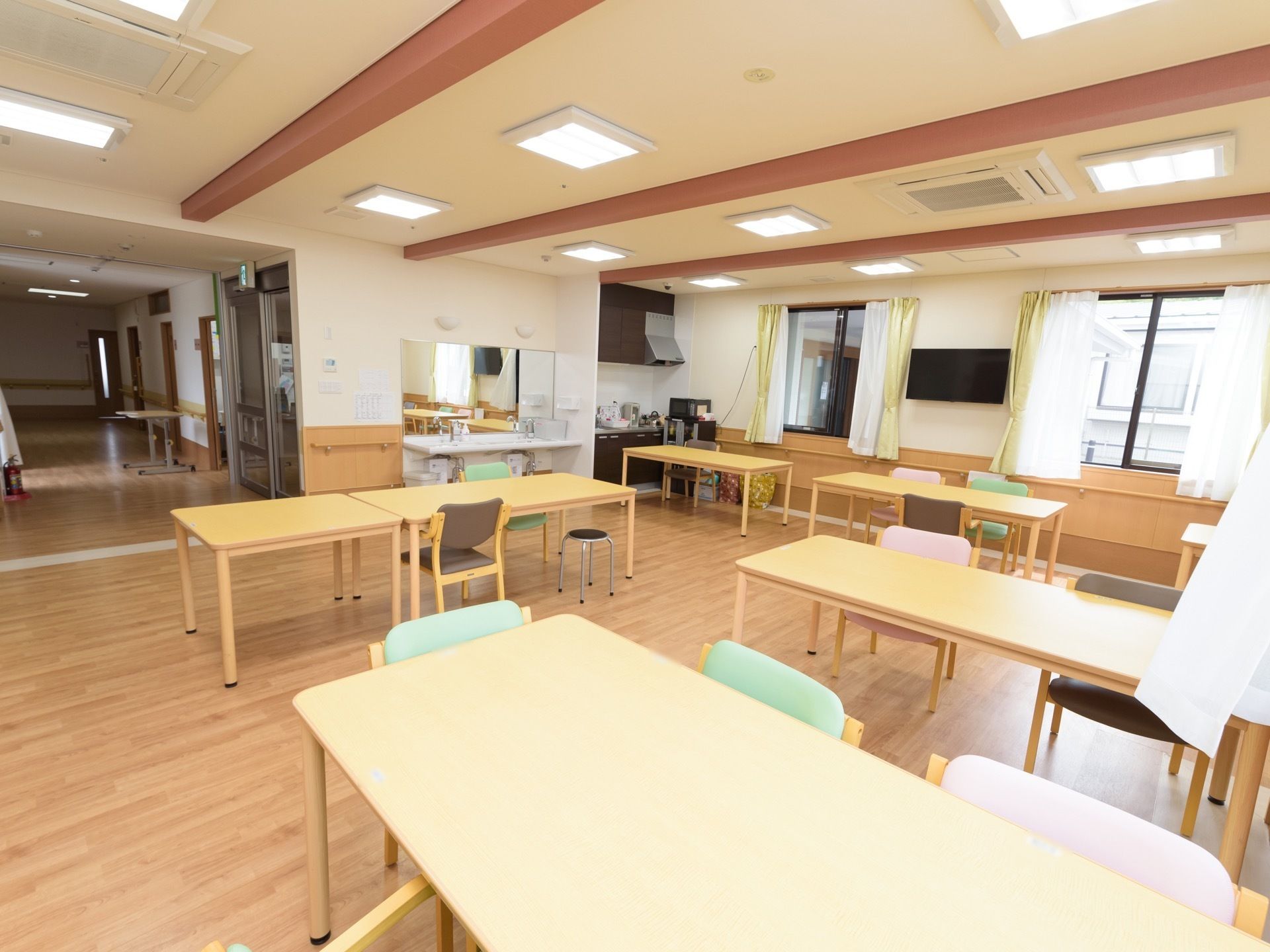 四角い照明器具が複数設置されている明るい空間に、テーブルや椅子が並べられている。左側に洗面スペースやキッチンがある。