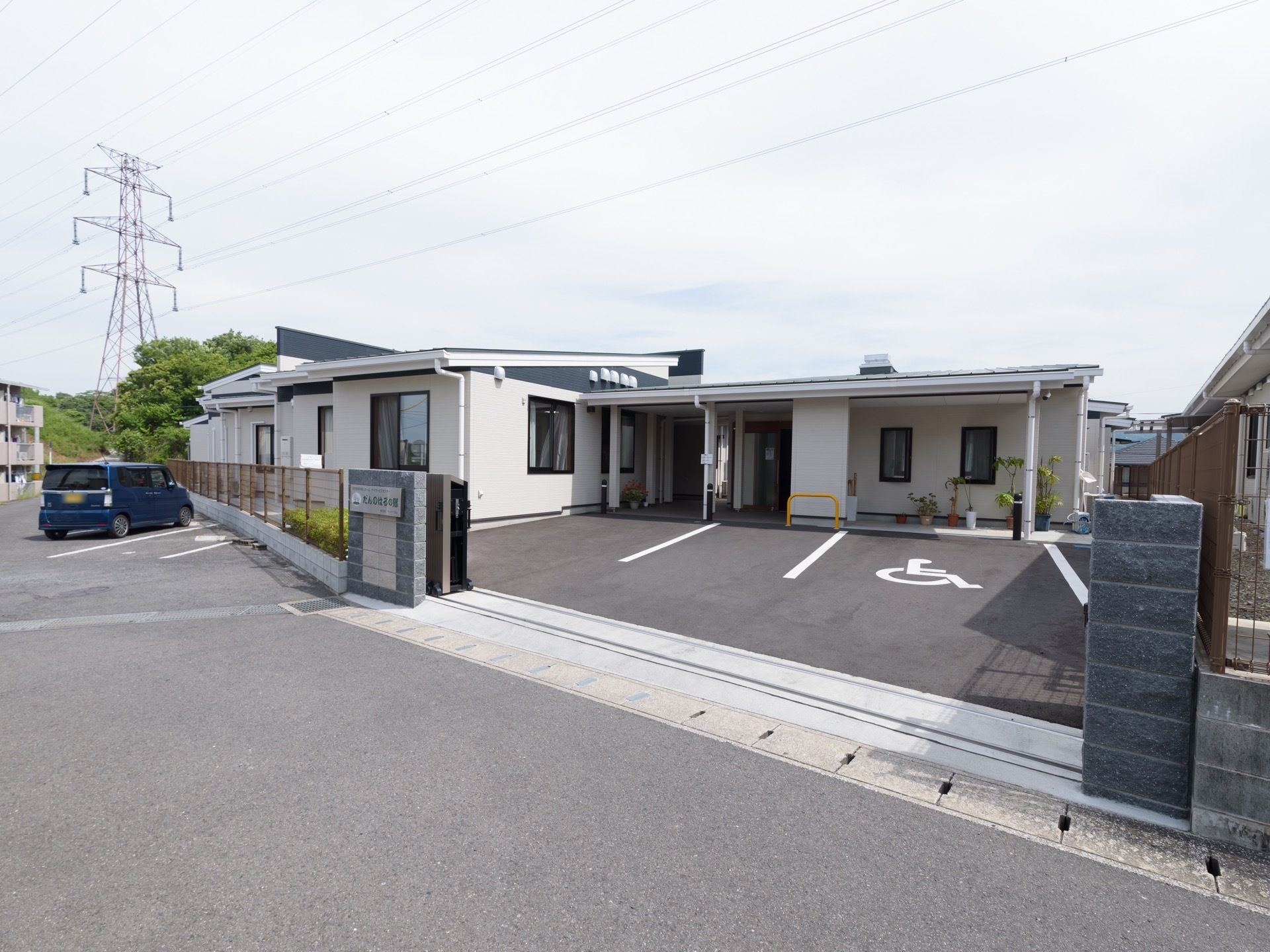 アスファルトで舗装された敷地に、平屋建ての施設が建っている。周囲には、集合住宅や緑豊かな木々が見える。