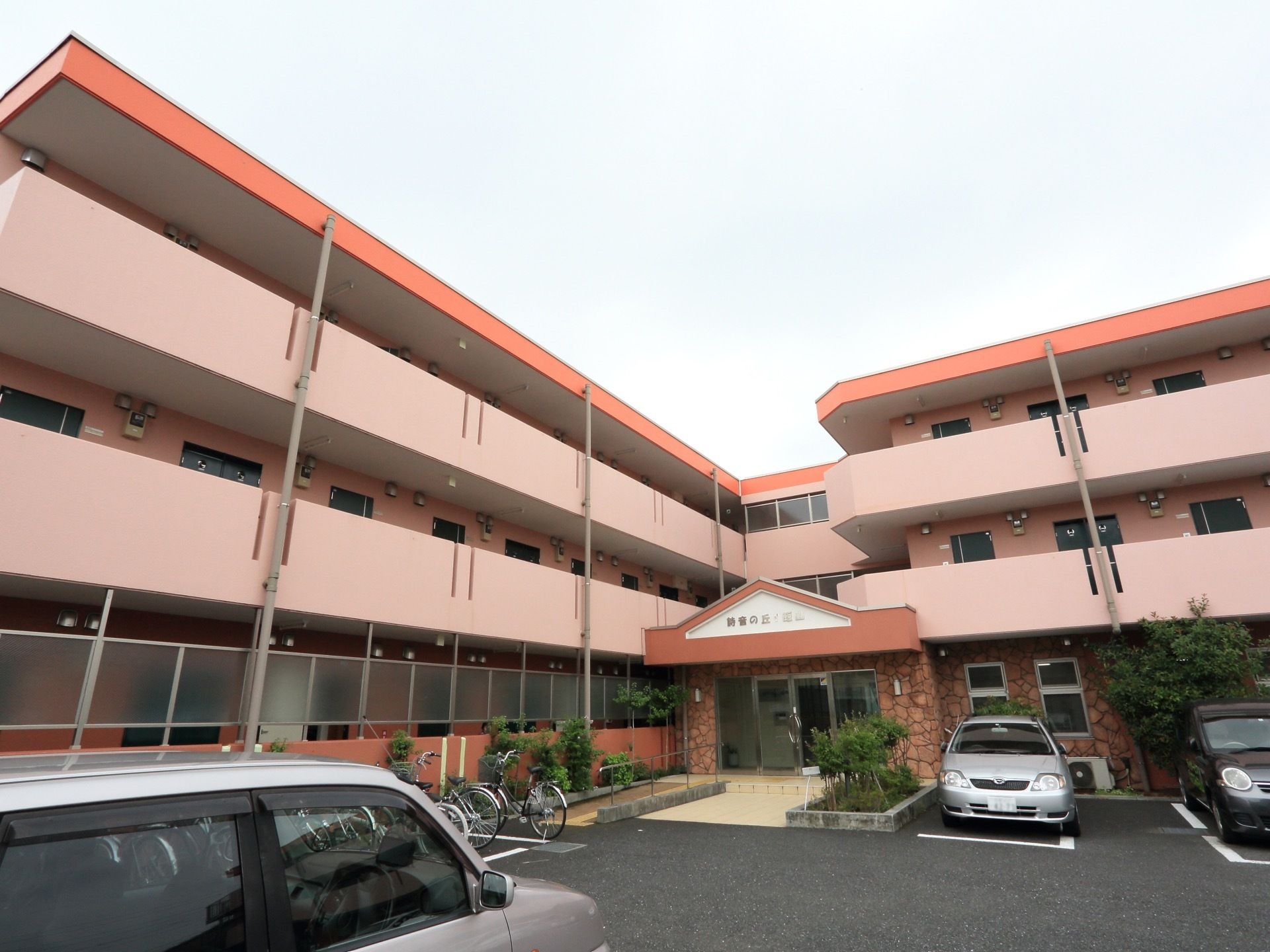 3階建てのピンク色の建物。１階部分はすりガラスの目隠しでプライバシーを確保している。駐車場と駐輪場があり、車や自転車が数台停まっている。