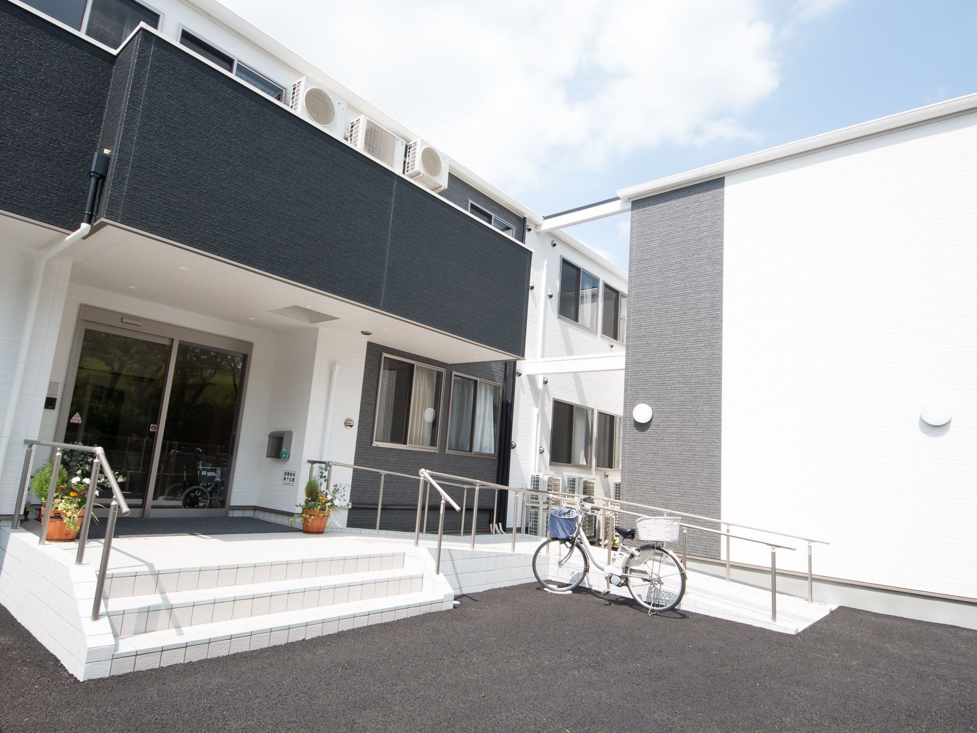 2階建ての施設で、外玄関付近の様子である。3段程の階段とスロープ、手すりが取りつけられている。玄関前の両側にお花が飾られている。