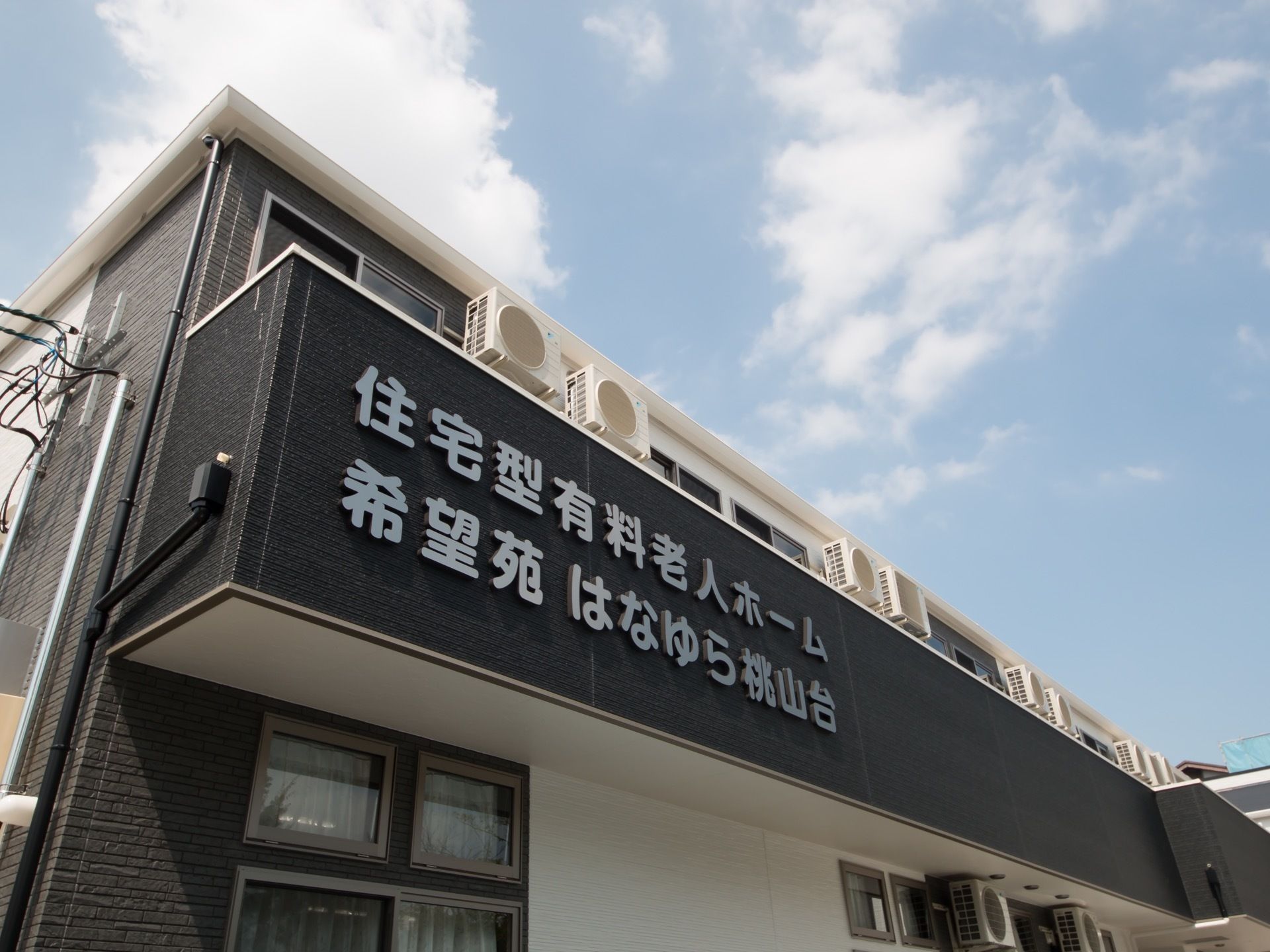日当たりの良い施設建物のベランダにホーム名が取りつけられている。窓が多くあり、室外機が置かれている。