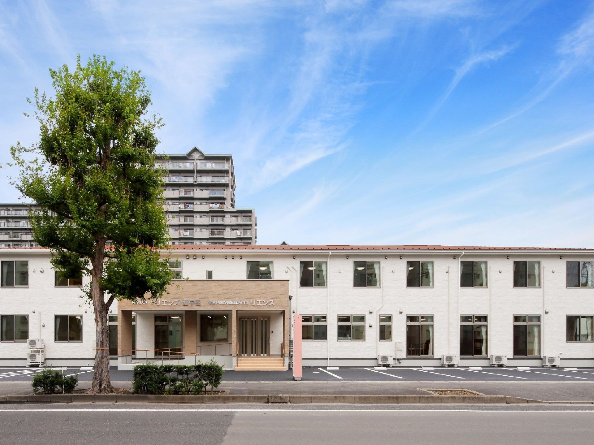 玄関正面から。２階建ての建物は全体的に白いが、屋根と玄関周りがベージュ。ホームの前には木が植えられている。