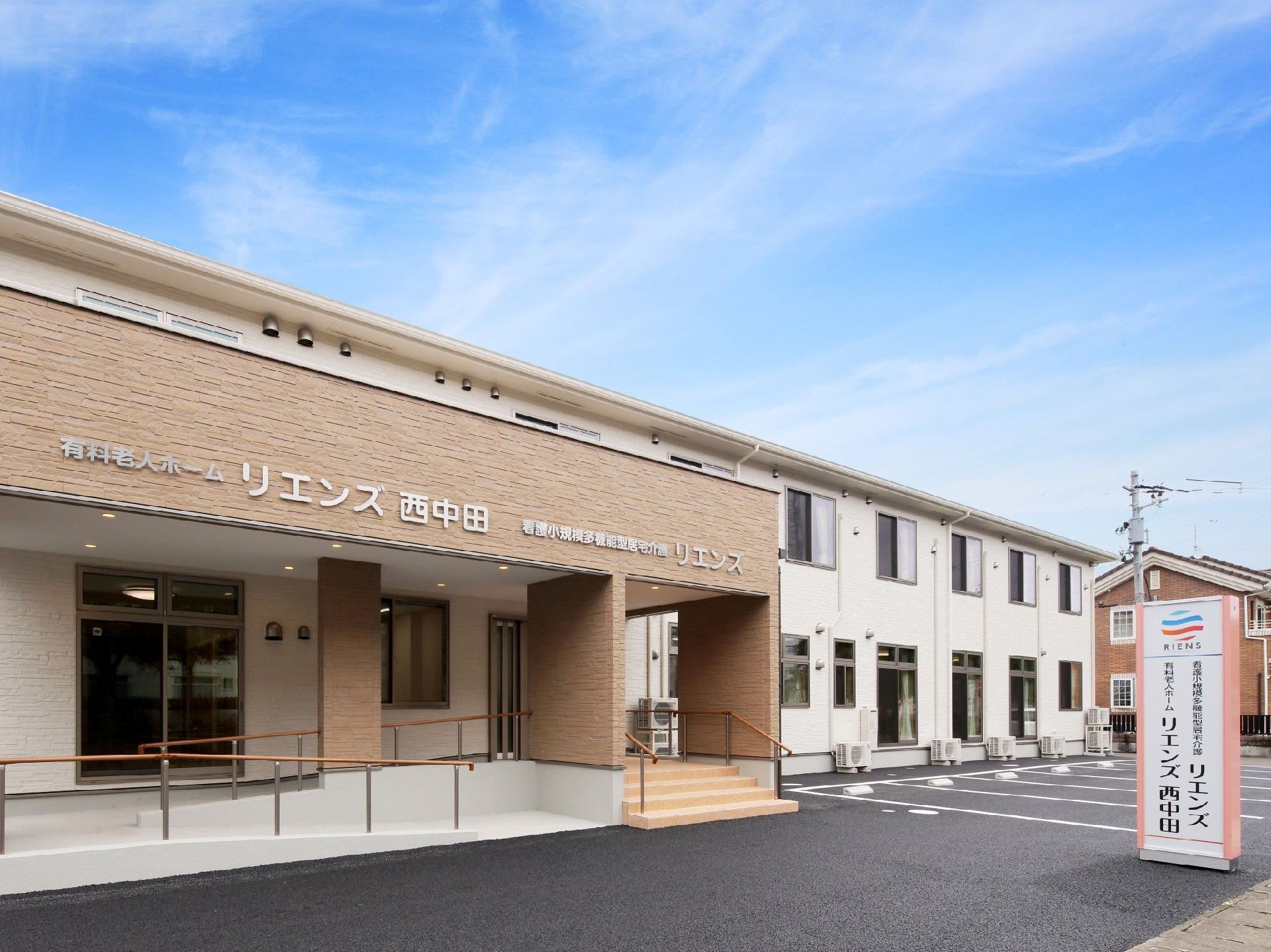 白い２階建ての建物。玄関周辺はほかの部分より前に出ていてベージュとなっている。玄関周りにはスロープも設置。