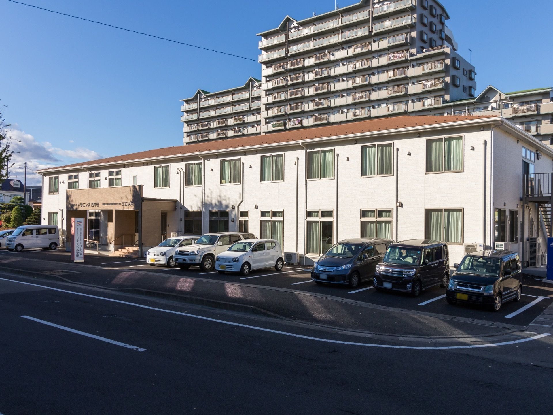 白い壁にオレンジの屋根の2階建ての施設建物である。建物の中央に玄関と、建物に沿った駐車スペースがある。