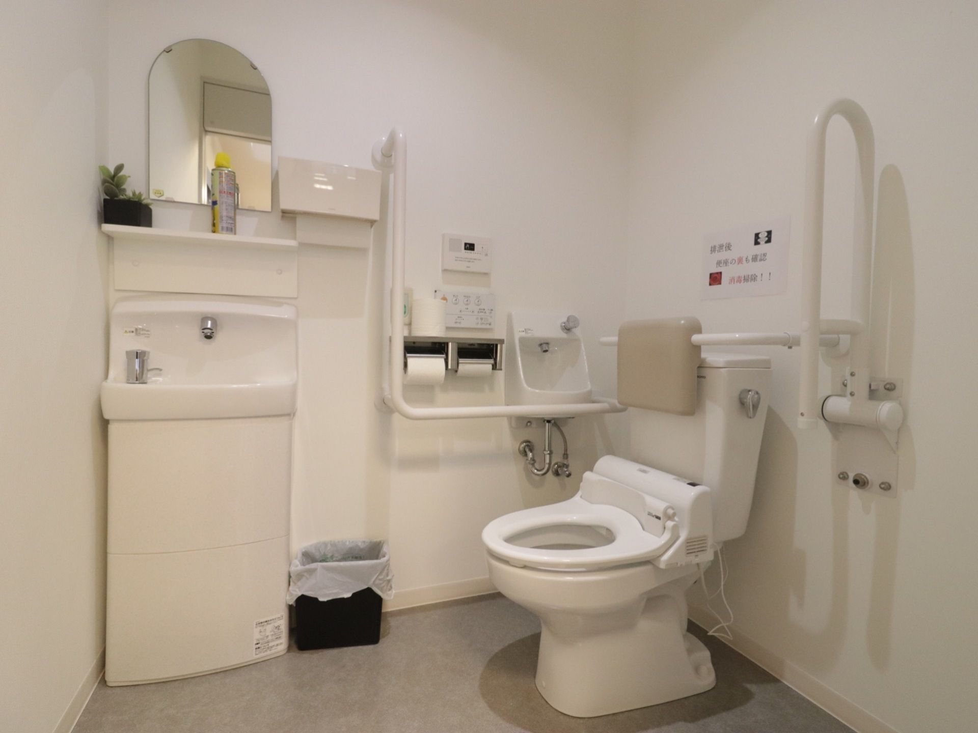 広くて清潔感のあるトイレである。洗面台と洗浄機能付きの便器が設置されている。便器の周りの壁には随所に手すりが取りつけられている。