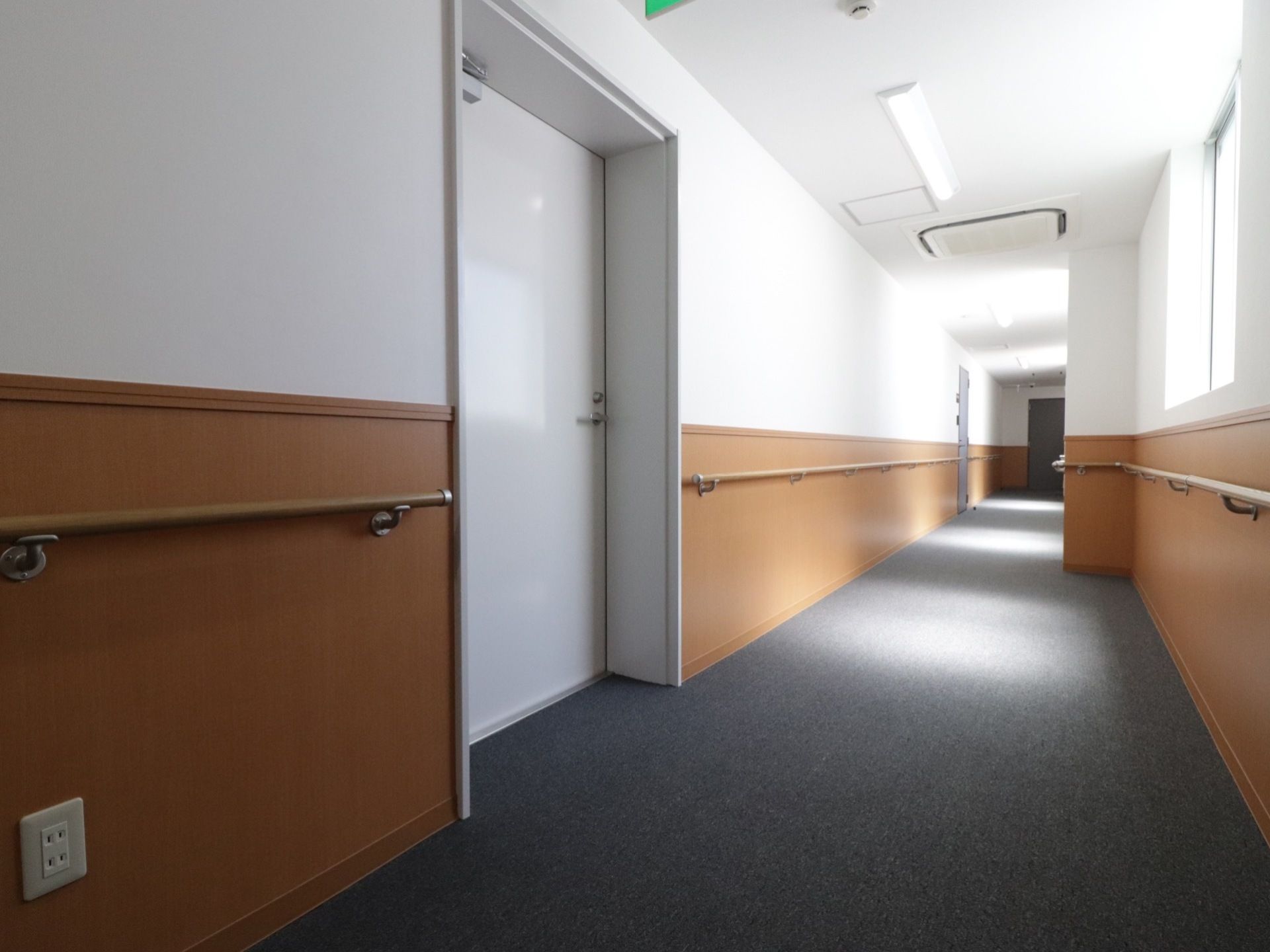 横幅の広い廊下である。両側の壁には手すりが設置されている。窓から日差しが差し込んでいる。廊下に面した部屋のドアは白く塗られている。