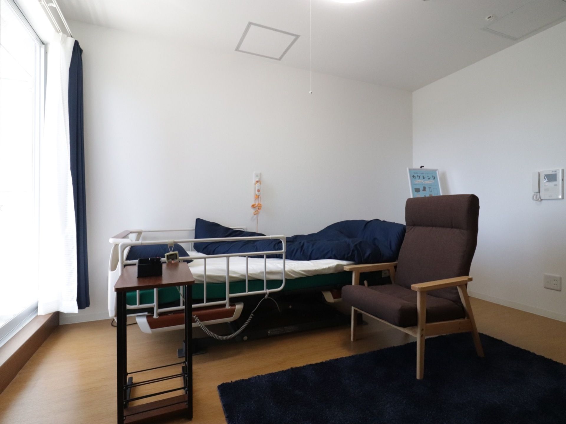 フローリング床の部屋に手すり付きのベッドが置かれている。ベッド脇にはひじ掛け付きの椅子がある。壁にナースコールが設置されている。