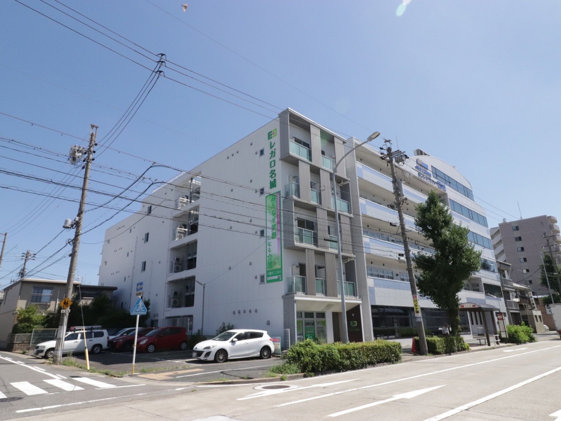 白い５階建ての建物である。建物の壁に大きく施設名が書かれている。施設の横は駐車場となっており、数台の車が停めてある。