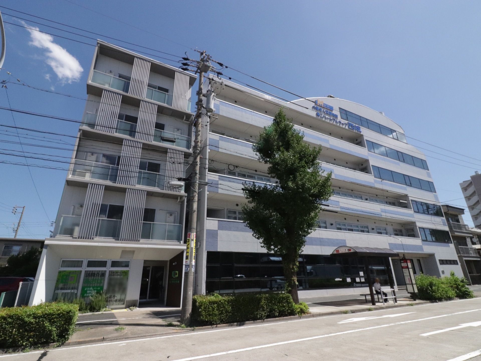 ホワイトとダークブラウンの5階建ての建物である。施設の前は歩道、車道があり、近くには、バス停がある。