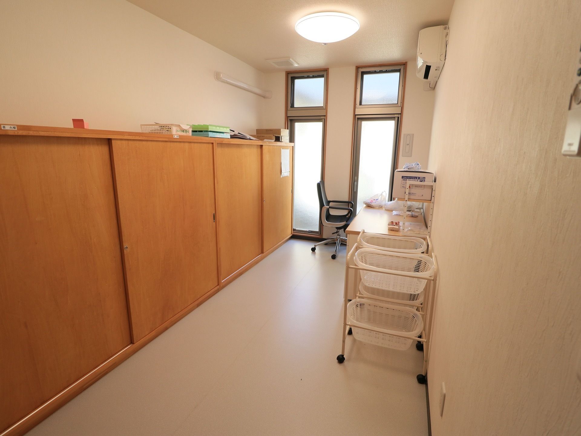 適度な空間の往診室である。引き戸式の棚が壁一面に設置され、先生用のテーブルと椅子、物置用のかごが置かれている。