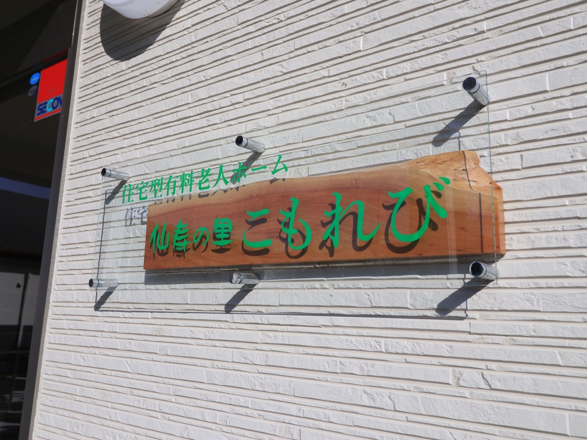 施設の壁に、緑色の施設名を記したクリアなプレートが設置されている。プレートの後ろに木板を置いてアクセントをつけている。