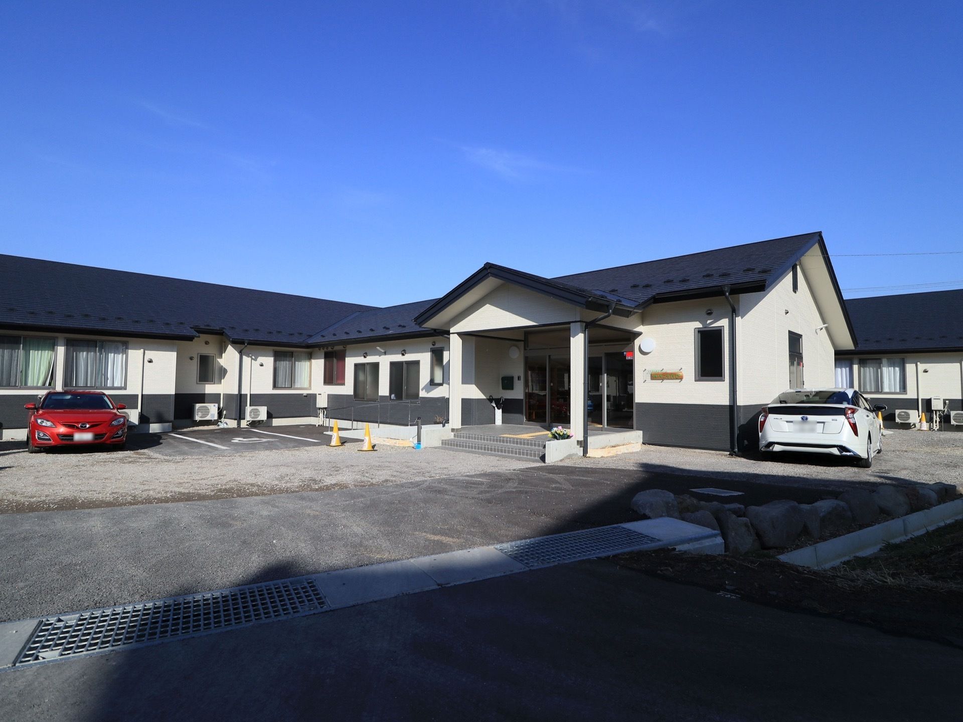 平屋建ての施設の前は、開放感のある駐車スペースとなっている。玄関へは、２段の階段の他、手すり付きのスロープも設置されている。