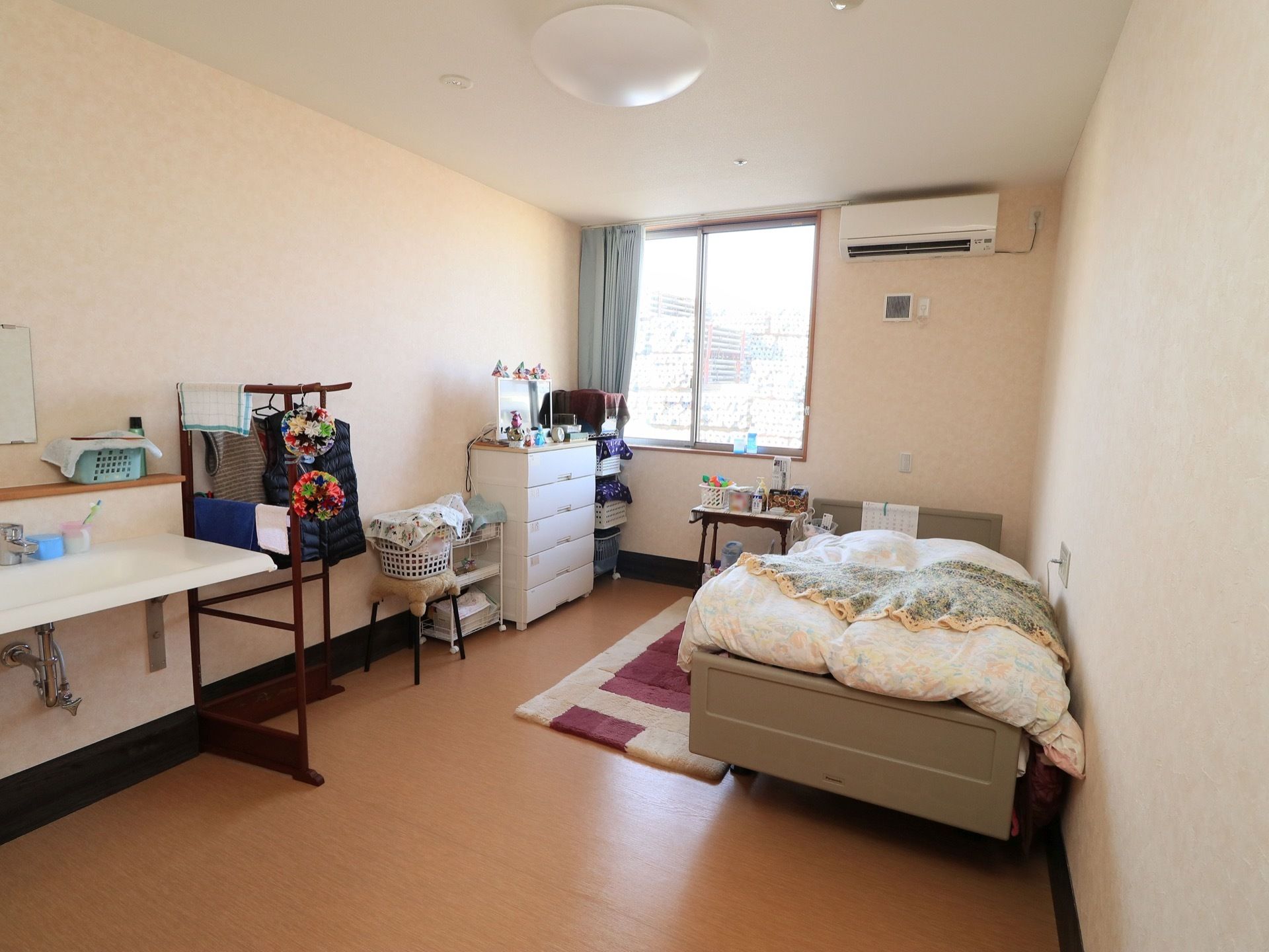 清潔感のある居室である。窓からは、十分な光が差し込んでいる。ベッドや家具、ハンガーなどが置かれている。