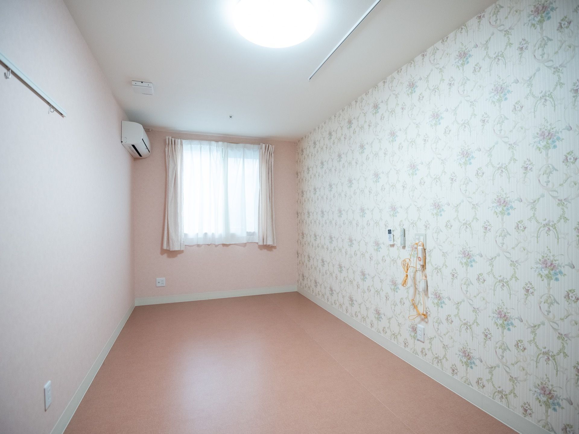 あたたかいピンク色の床に施工された居室。エレガントな花柄の壁紙が印象的である。窓のそばには、エアコンが備えられている。