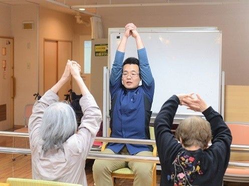 ブルーのユニフォームを着た男性スタッフが、女性入居者２名に対して体操の指導をしている。３人ともいすに座り手を上に伸ばしている様子。