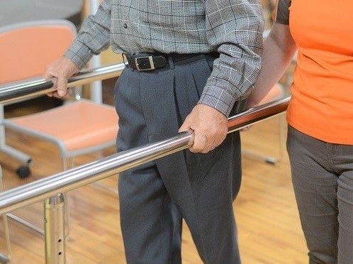 平行棒を握り歩行訓練を行う男性と腰を支え介助するスタッフ。