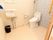 クリーム色のタイルが貼られた床に白いタンク付きの洋式トイレがある。洗面台の上に鏡が設置され、その下に黒いゴミ箱が置いてある。