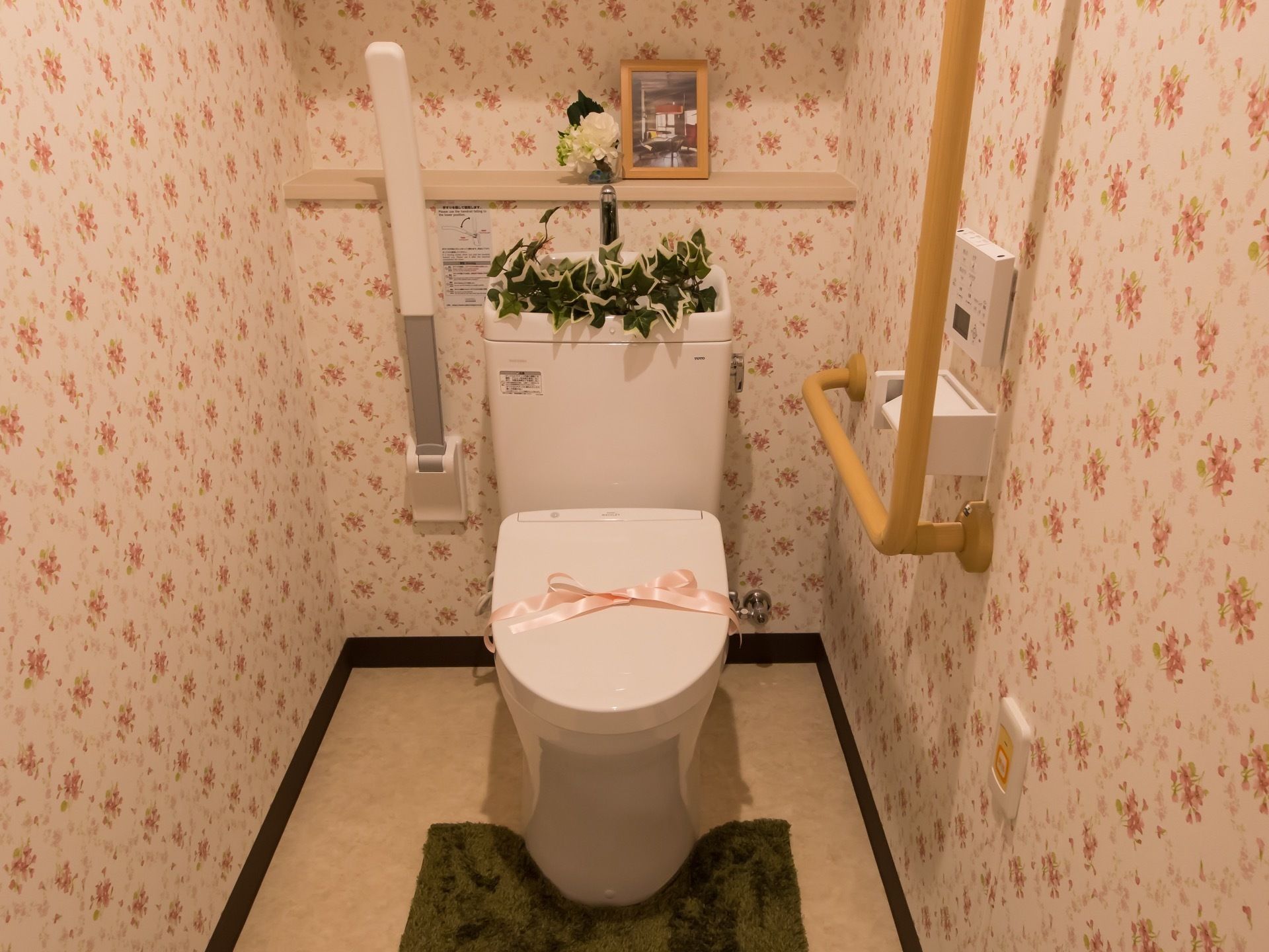 花柄の壁紙で囲まれた洋式トイレがある。便座の横には可動式の手すりがあり、壁にはナースコールが付いている。