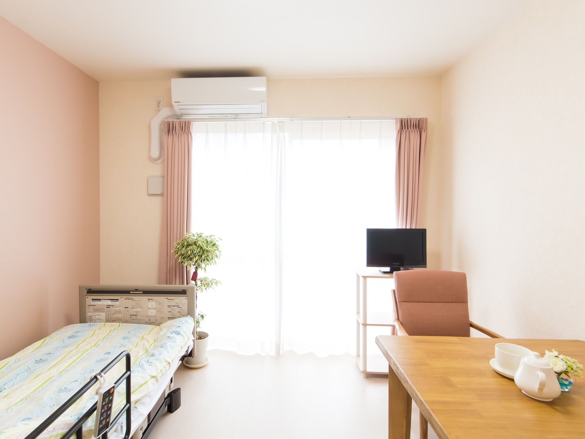 掃き出し窓のある明るい居室に、電動ベッドとダイニングテーブル、テレビが置かれている。窓の上にはエアコンがある。