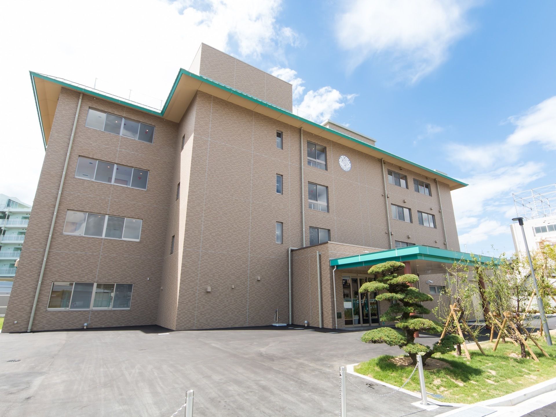 舗装された敷地に４階建ての茶色いビルが建っている。植栽があり、松の木などが植えられている。
