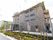 植栽があり、奥に４階建ての住宅が建っている。自動ドアの玄関の他、建物の脇には非常用の外階段も設置されている。