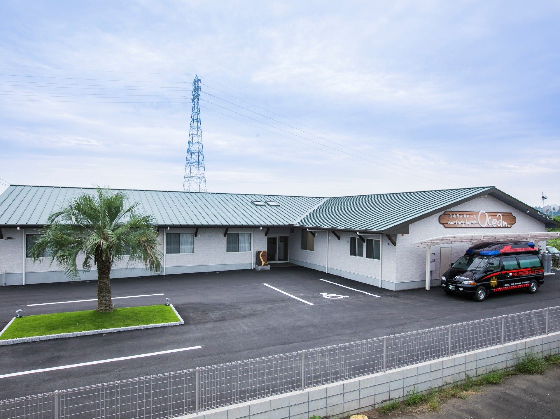 平屋建ての建物はエル字型で外壁が白、屋根が緑。敷地内には黒と赤の車が一台止まっている。