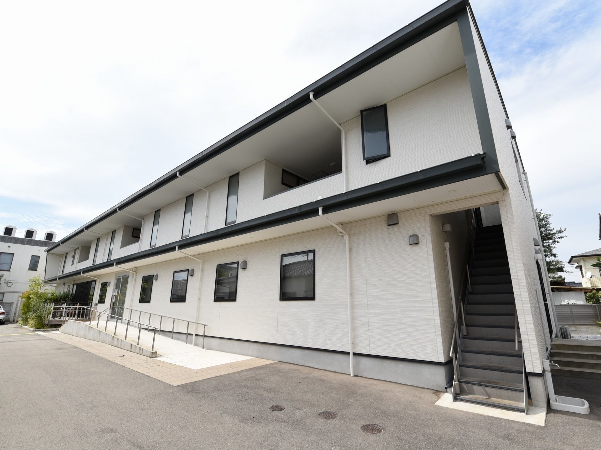 施設は白色の外壁で、2階建ての建物となっている。右側には階段があり、左側にはスロープが設けられている。