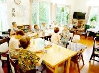 共有スペースに集まる入居者。窓が大きく明るい雰囲気。奥にはテレビや観葉植物が置かれている。