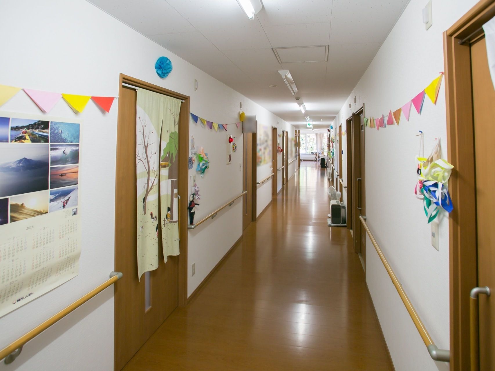 廊下の壁には手すりが両側に備わっている。さらに、入居者様手作りの作品が飾られ、カレンダーが掛けられている。