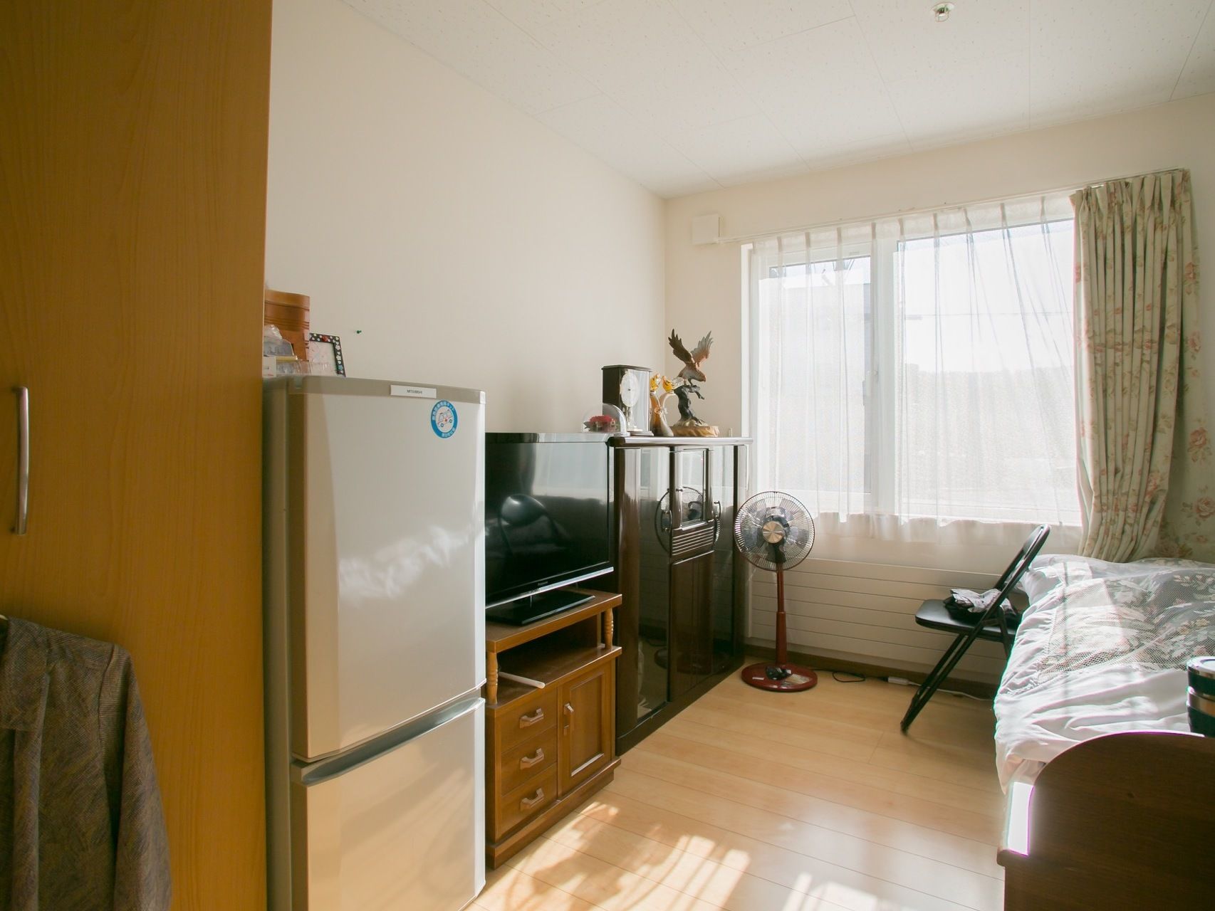 フローリングの部屋に、冷蔵庫とテレビ台に乗ったテレビ、木製の棚が置かれている。反対側にはベッドがあり、床には扇風機がある。